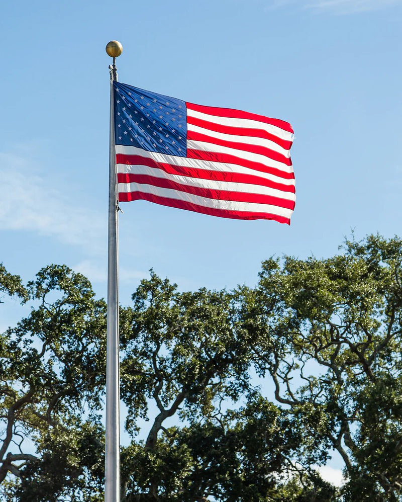 Load image into Gallery viewer, A bright aluminum flagpole with an external halyard flying a large American flag fully extended against a blue sky above green trees.