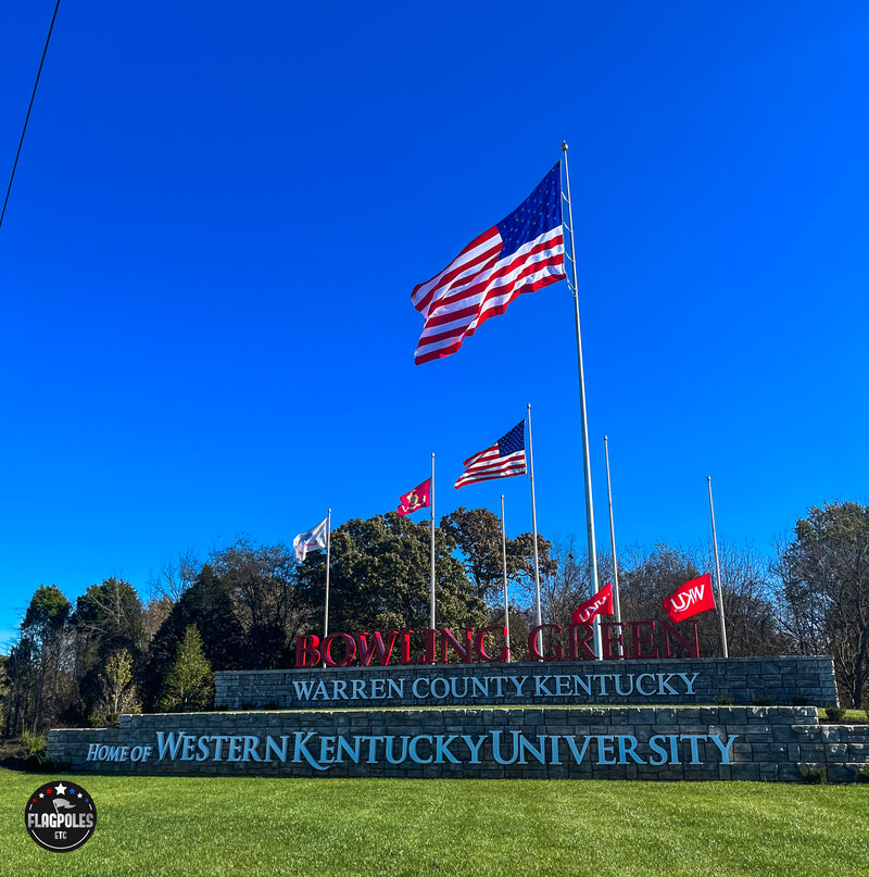 Load image into Gallery viewer, A tall aluminum flagpole with an external halyard system displaying the American flag, set against a clear blue sky, with greenery and a sign for Western Kentucky University in the foreground.