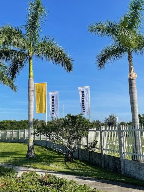 Load image into Gallery viewer, A roadside view of three fiberglass banner poles with vertical banners, installed behind a fence and framed by palm trees.