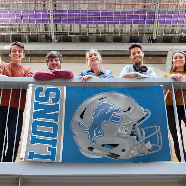 Load image into Gallery viewer, Detroit Lions Helmet Flag displayed on a railing with five college-aged fans standing behind it, showing the flag's large size and vibrant silver and blue helmet graphic in use.