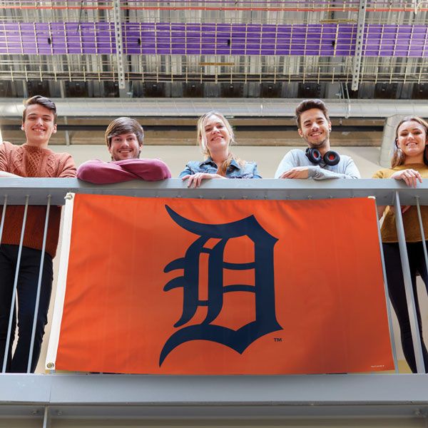 Load image into Gallery viewer, Detroit Tigers Deluxe Flag displayed on a railing, held by five smiling fans, showcasing the large navy 'D' logo against the orange background in a stadium or campus setting.