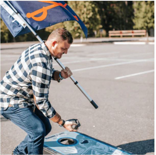 Load image into Gallery viewer, A man shown tailgating and playing cornhole while holding the Hide-A-Pole portable flagpole with a flag attached. The image demonstrates the pole's use as a lightweight, quick-setup outdoor display for events like tailgates or camping.