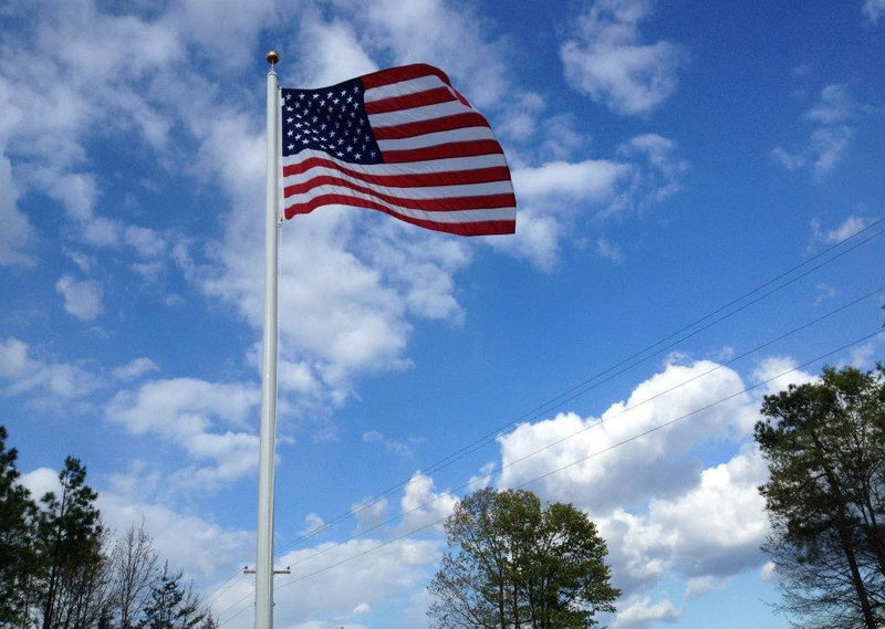 Load image into Gallery viewer, 65-foot Lincoln series flagpole viewed from below with large US flag and bright blue sky as a backdrop. A few trees are also visible.
