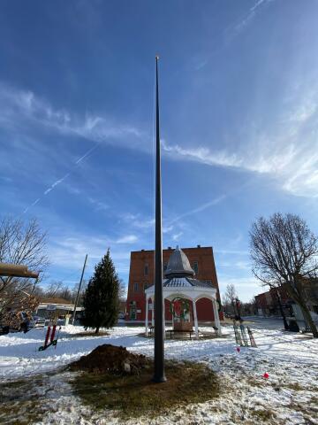 75-foot Lincoln series flagpole, installed in center of park pavilion with gazebo and brick building visible in the background, no flag is attached.