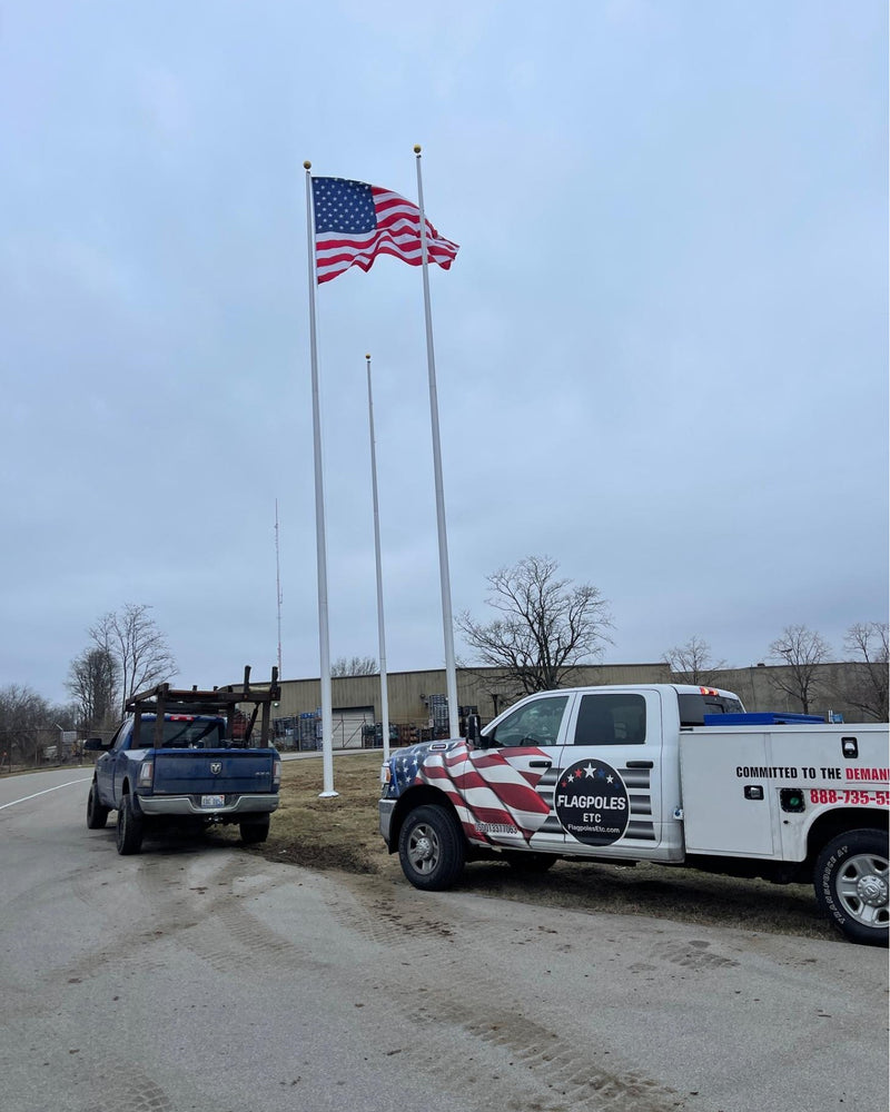 Load image into Gallery viewer, An aluminum flagpole with a traditional top mounted on a base, with an American flag displayed at the top, installed beside a road with vehicles in the background.
