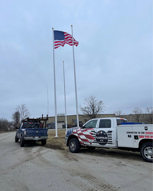 An aluminum flagpole with a traditional top mounted on a base, with an American flag displayed at the top, installed beside a road with vehicles in the background.