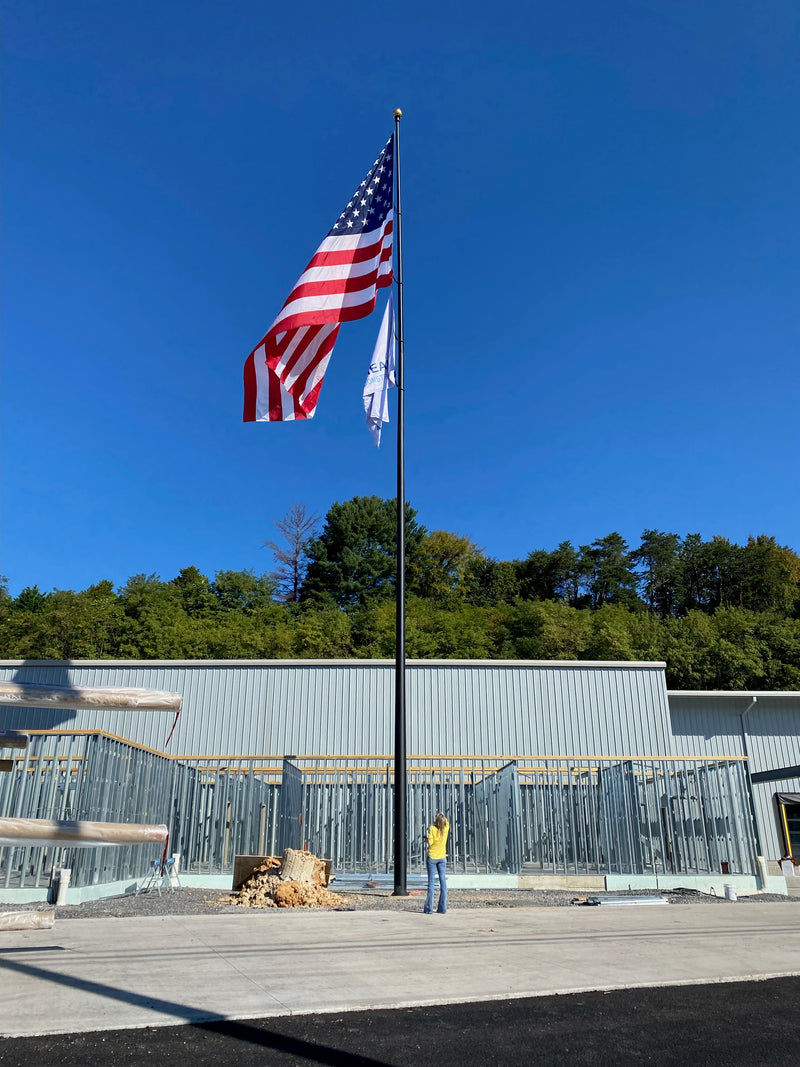 Load image into Gallery viewer, An aluminum flagpole with a reinforced welded door frame, installed in front of a building with a United States flag attached at the top, set against a clear blue sky backdrop.
