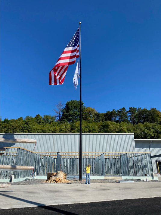 An aluminum flagpole with a reinforced welded door frame, installed in front of a building with a United States flag attached at the top, set against a clear blue sky backdrop.