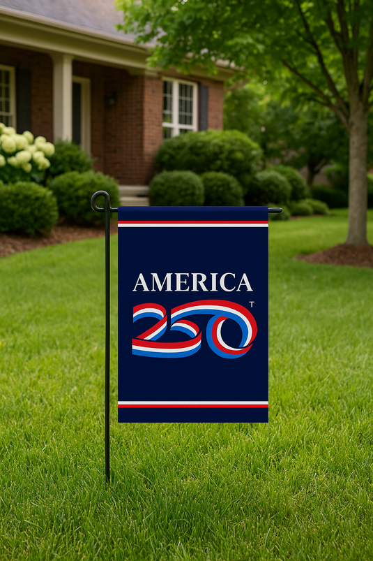America 250 garden flag mounted on a black metal stand in a neatly trimmed front yard, with a brick house and green trees in the background.