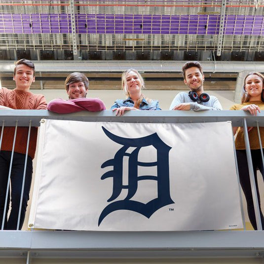 Detroit Tigers White Background Flag displayed on a railing, held by five smiling fans, showcasing the large navy 'D' logo against the white field in a stadium or campus setting.