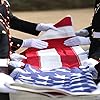 U.S. Memorial Flag being formally folded by uniformed military personnel wearing white gloves during a solemn burial or flag-draping ceremony. The flag is partially folded into the ceremonial triangle shape.