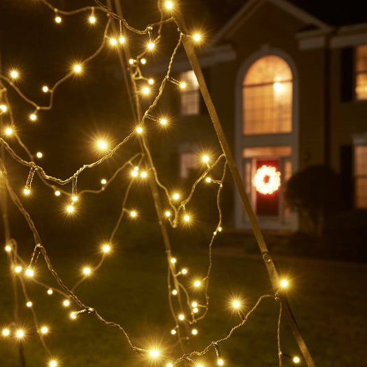 Close-up of the twinkling Fairybell Luminous Switch Flagpole Christmas Tree Light in warm white, with a house visible in the background.