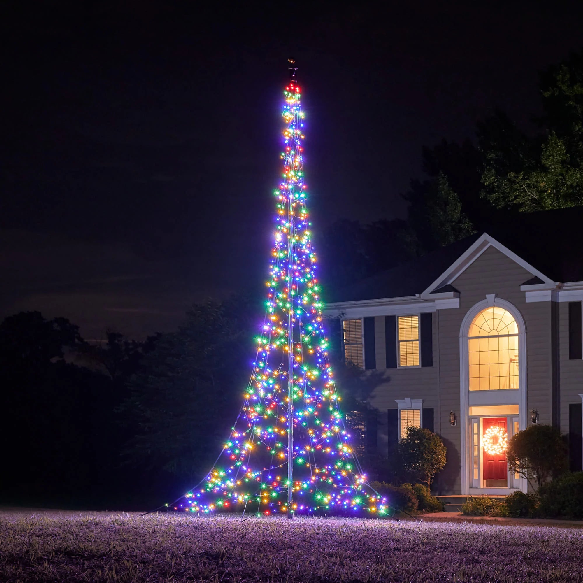 Large, multi-color illuminated Fairybell Flagpole Christmas Tree Light installed in a yard for holiday display at dusk.
