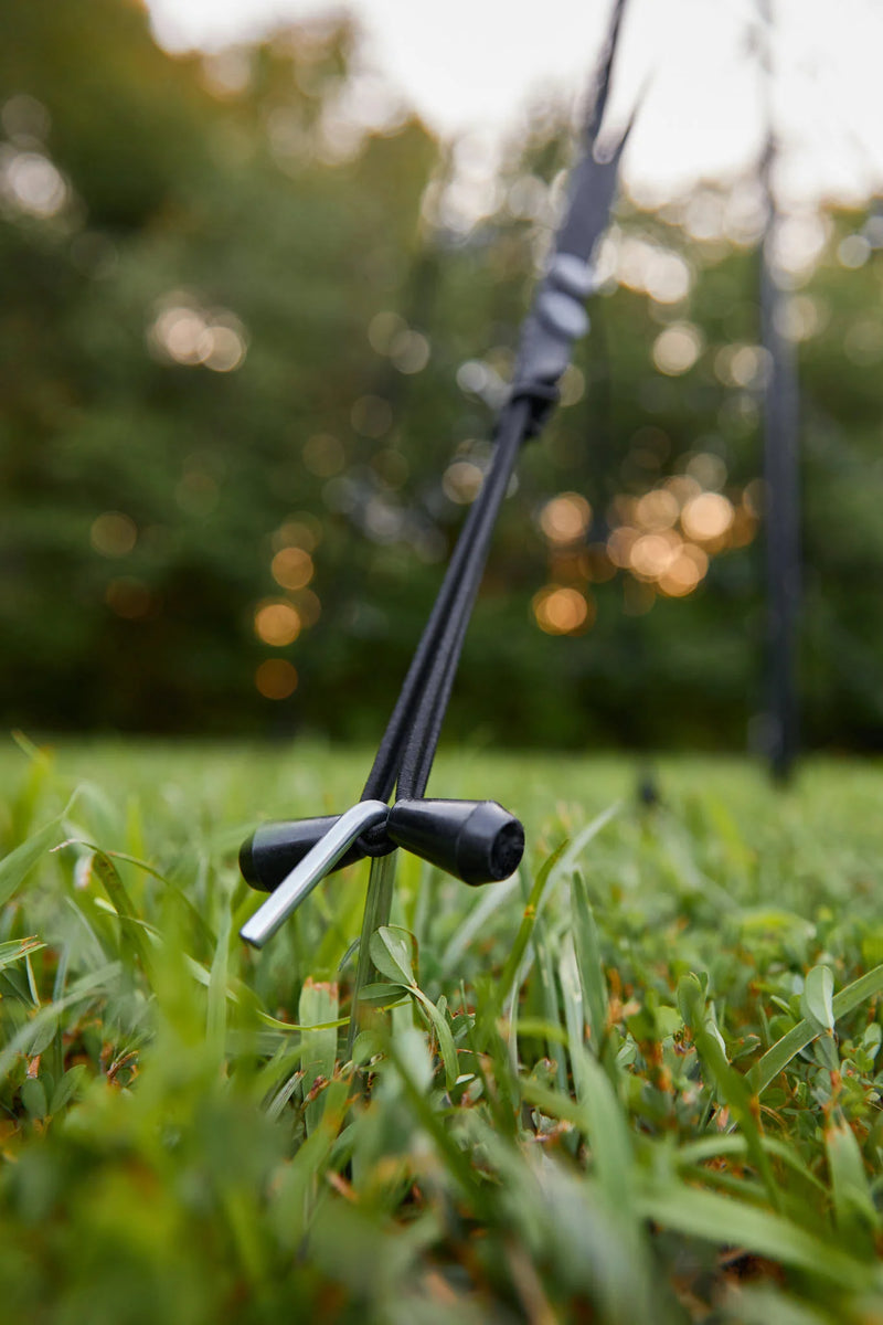 Load image into Gallery viewer, Close-up of the black tether and ground stake securing the Fairybell Single Color Flagpole Christmas Tree Light base in the grass.
