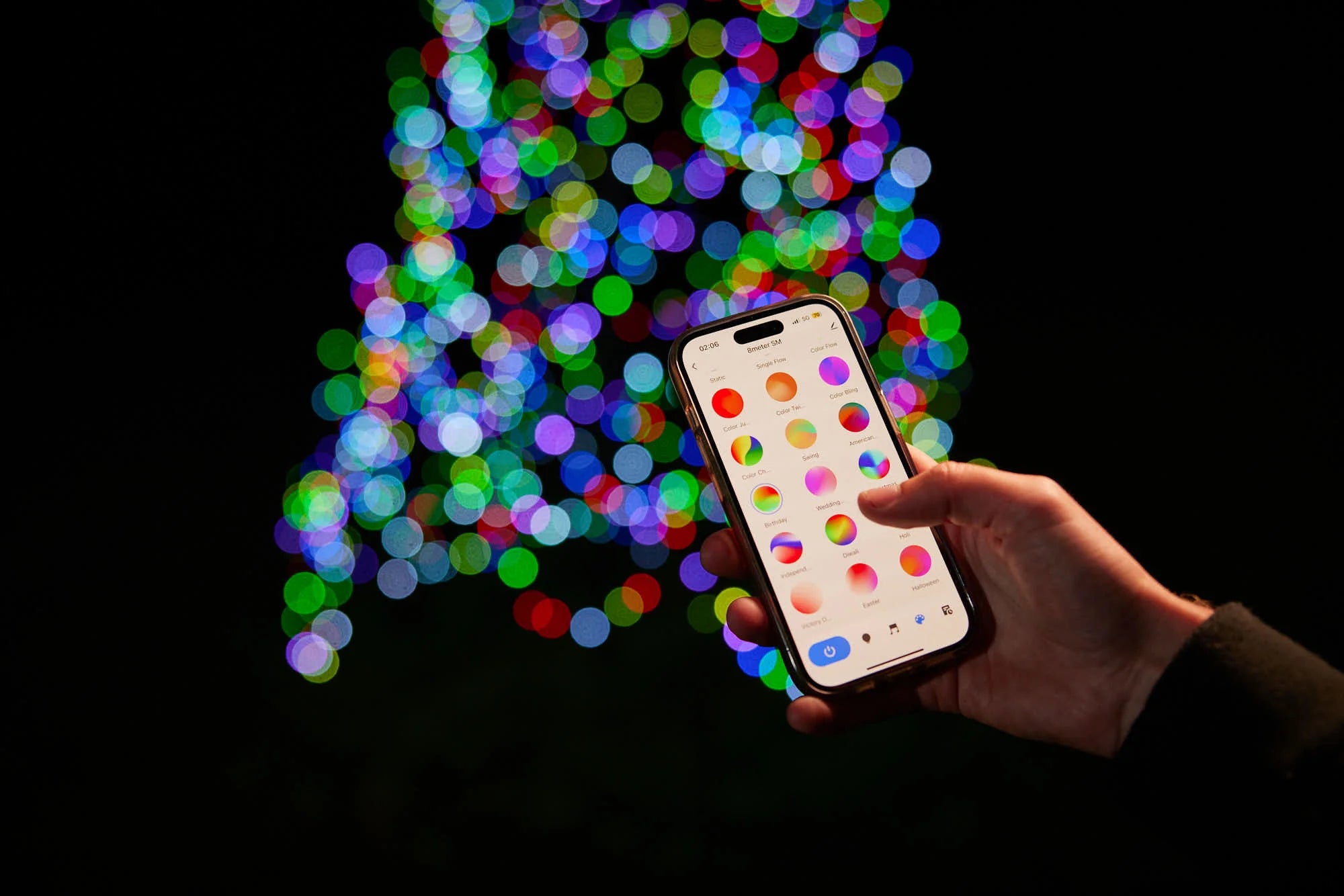 Hand holding a phone displaying the control app for the Fairybell Smart Magic Light against a blurred multicolor light background.