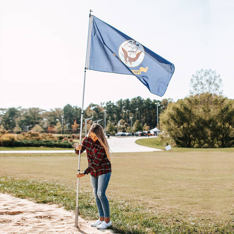 Load image into Gallery viewer, Woman standing outdoors setting up the Flagpole-To-Go Portable Telescoping Aluminum Flagpole which is flying a U.S. Navy flag, demonstrating the pole's use in a lawn or beach setting.
