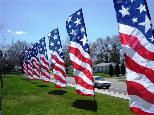 Row of vertical, feather-style US flags displayed outdoors along a suburban road, illustrating the use of the Flexible Rod for Super Pole that holds the top edge of the banner taut for maximum visibility even without wind.