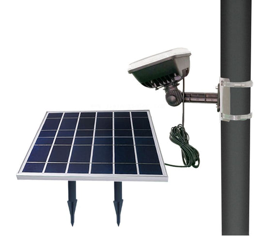 Close-up of the FPL Independence Solar Flagpole Light system mounted on a dark pole. The setup includes the large, flat solar panel (shown separate with ground stakes) connected by a black wire to the weatherproof LED floodlight unit, which is secured to the pole with clear mounting straps.