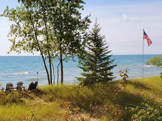 A Homesteader sectional aluminum flagpole proudly displaying an American flag near a lakeshore or beach. The pole is installed on a grassy dune, positioned near large trees, emphasizing its suitability for durable residential outdoor use in scenic or coastal environments.