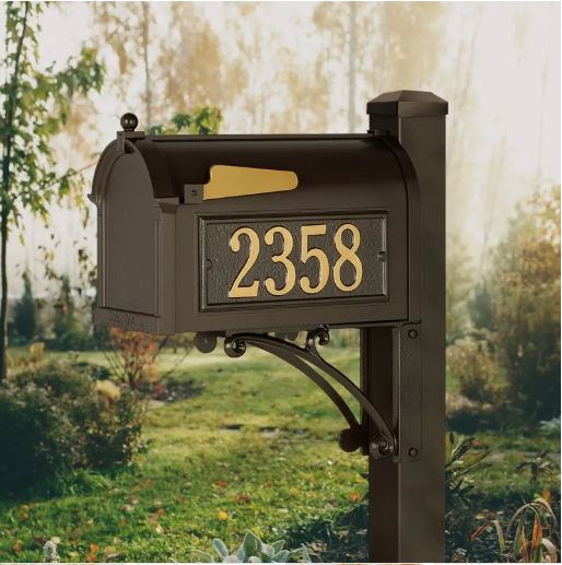Superior package-sized bronze mailbox installed on a matching post against an outdoor, wooded background. The mailbox features large gold address numbers (2358) and a curved black aluminum support bracket.