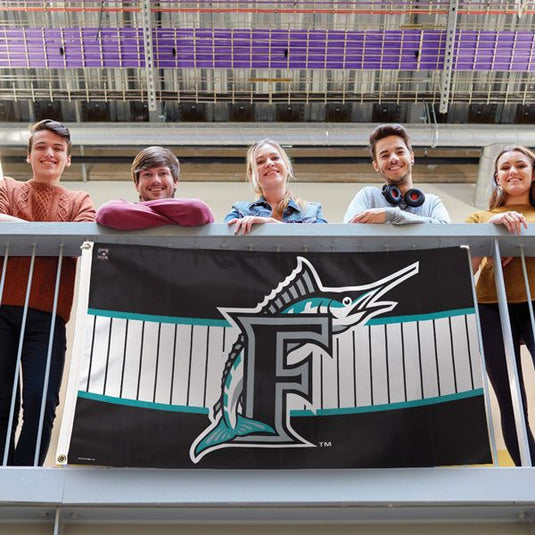 Five young fans hold the Miami Marlins Cooperstown 3x5 MLB sport flag over a balcony railing. The black flag features a teal-striped design and the team's marlin logo over a bold 'F'.