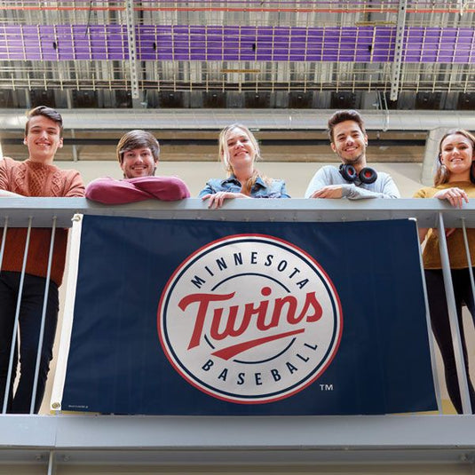 Lifestyle shot of the 3x5 Minnesota Twins flag with its navy blue and white circular logo being held and displayed over a balcony railing by a diverse group of five smiling young adults.