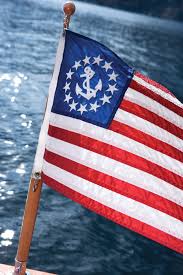Load image into Gallery viewer, Close view of the U.S. Yacht Ensign Flag flying from a wooden staff over sparkling blue water.
