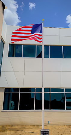 A tall aluminum flagpole with an American flag flying at the top, located in front of a modern building with large windows.